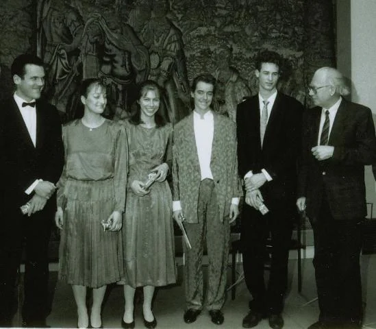 Schwarzweißfoto von sechs Personen in Konzertkleidung, die nebeneinander vor einem großen Wandteppich mit historischer Bildszene stehen (Uraufführungsinterpreten von Bertold Hummels op. 89a)