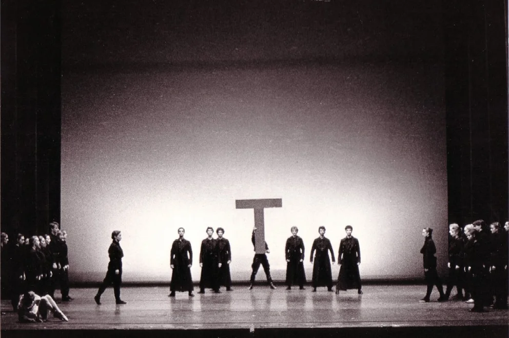 Black-and-white photograph of a ballet scene: Dancers in dark coats stand in two groups facing each other on an illuminated stage; in the centre is a figure with an oversized letter ‘T’, from ‘The Last Flower’ (music: Bertold Hummel, Op. 55a)