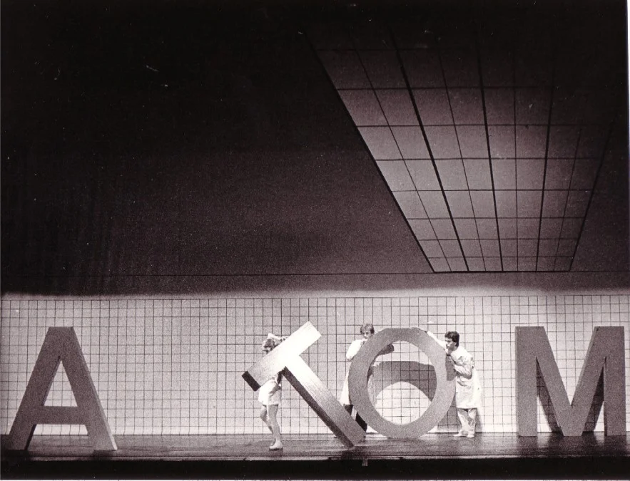 Black-and-white photograph of a ballet scene against a tiled backdrop: Dancers in white costumes move large letter sculptures that spell out the word ‘ATOM’; a still from the 1984 Augsburg performance of ‘Die letzte Blume’ (music: Bertold Hummel, Op. 55a)