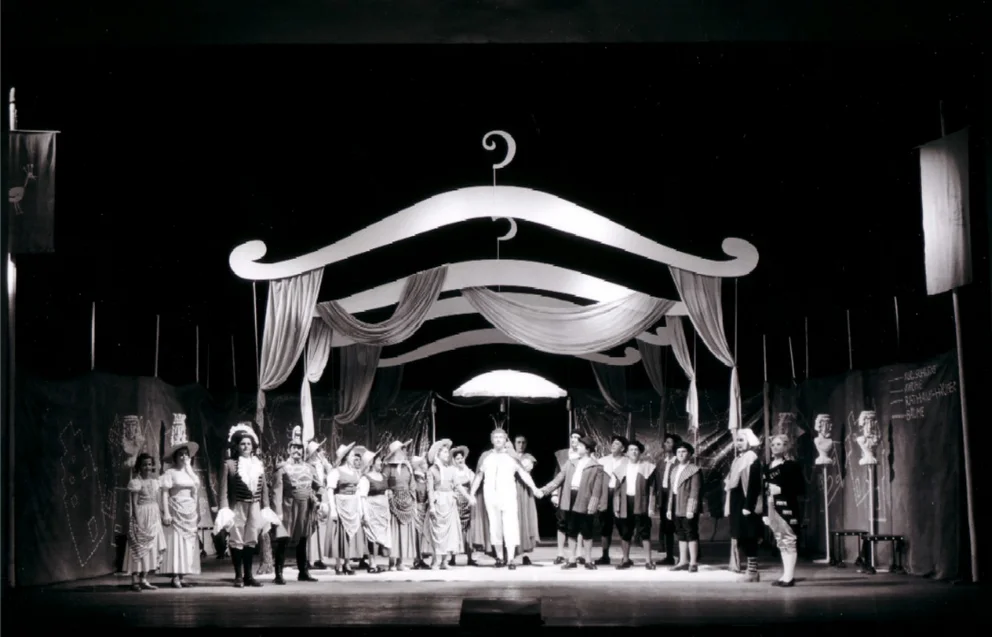 Photo de scène en noir et blanc prise lors de la création de l’opéra de chambre de Bertold Hummel « Les habits neufs de l’empereur », op. 10 (19 juin 1957, Fribourg) : grande scène d’ensemble, l’empereur, vêtu de ce qu’il croit être son nouveau costume, avance sous un dais à travers la foule des courtisans et des citoyens (5e image)