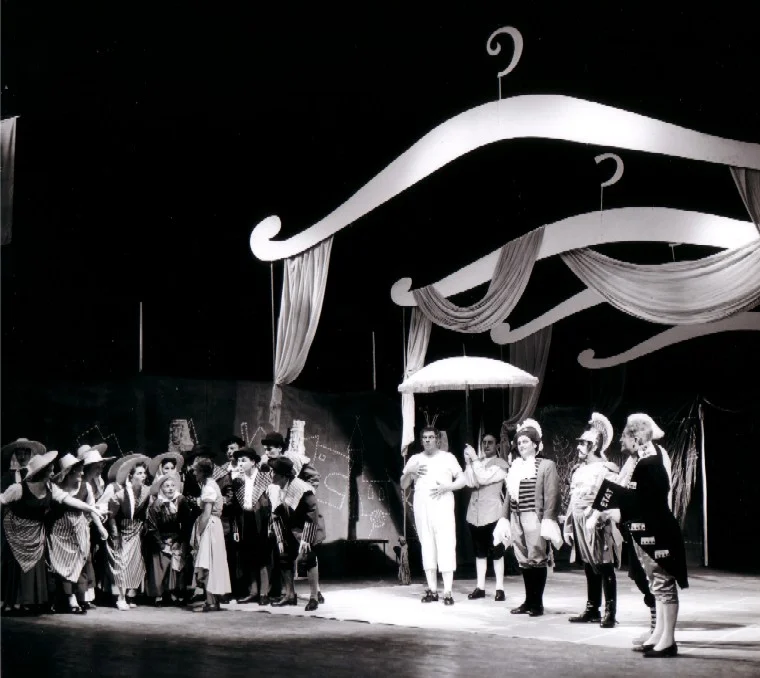 Photo en noir et blanc prise lors de la création de l’opéra de chambre de Bertold Hummel « Les habits neufs de l’empereur », op. 10 (19 juin 1957, Fribourg) : l'empereur avance sous un petit dais, entouré de courtisans et d'une foule qui l'attend (3e image)