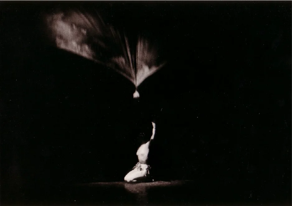 A dark black-and-white photograph of a single dancer kneeling in the spotlight, with a bright, fan-shaped beam of light above; a still from the production ‘Die letzte Blume’ (Music: Bertold Hummel, Op. 55a), Würzburg 1975