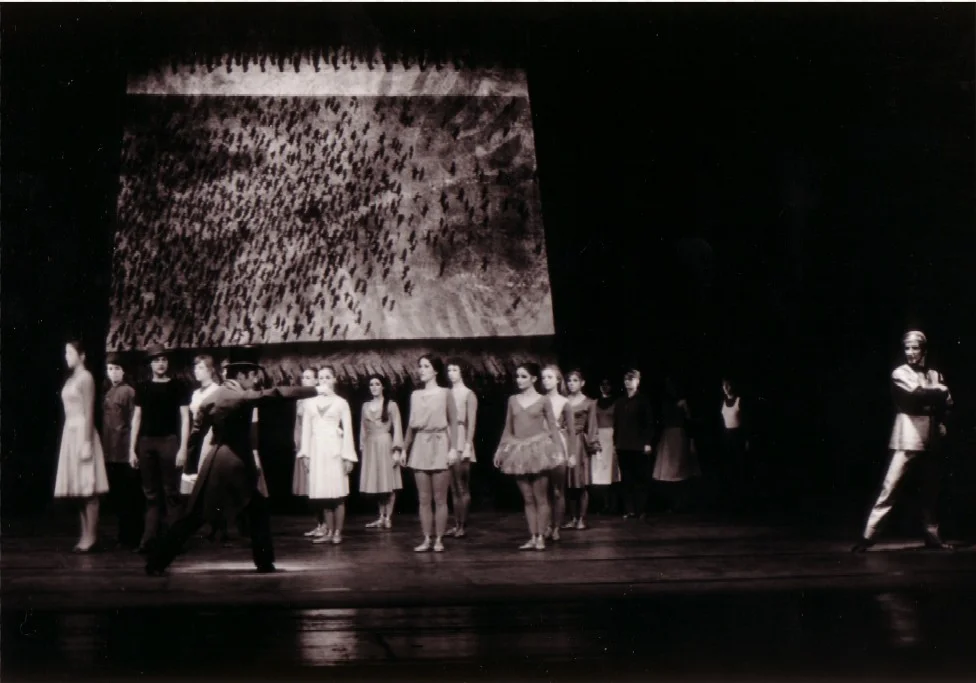 Black-and-white photograph of a ballet scene: a group of dancers in everyday clothes stand in front of a backdrop featuring an aerial view of a field; a figure in a coat and hat strides forwards with an outstretched arm. Scene from ‘The Last Flower’ (Music: Bertold Hummel, Op. 55a), Würzburg 1975