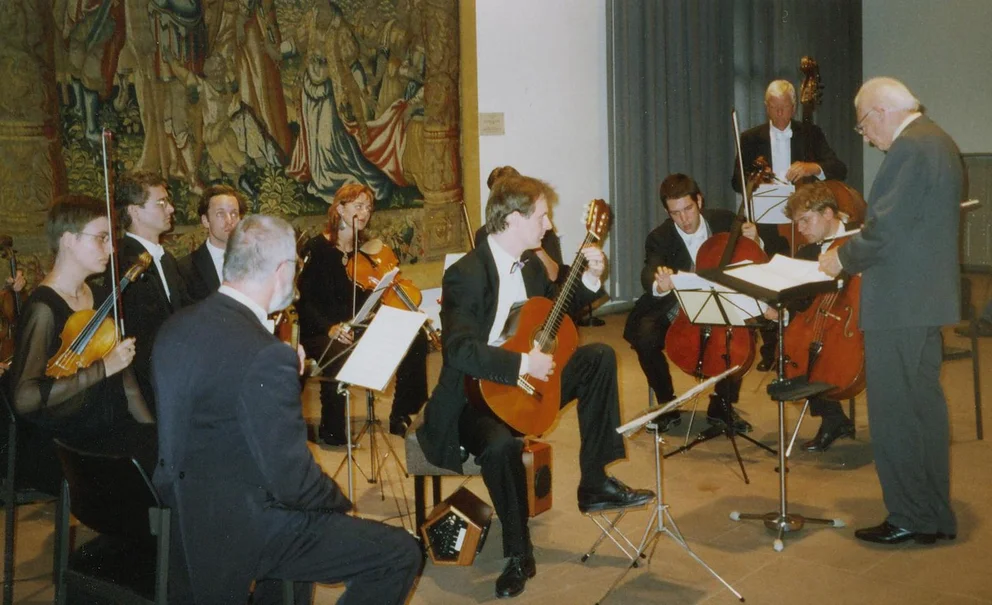 Photo couleur d'un concert d'orchestre de chambre : un guitariste est assis au centre devant les cordes (violons, violoncelles, contrebasse), un chef d'orchestre d'un certain âge se tient debout au premier plan devant son pupitre, avec en arrière-plan une grande tapisserie historique (création de l'œuvre op. 89b de Bertold Hummel)