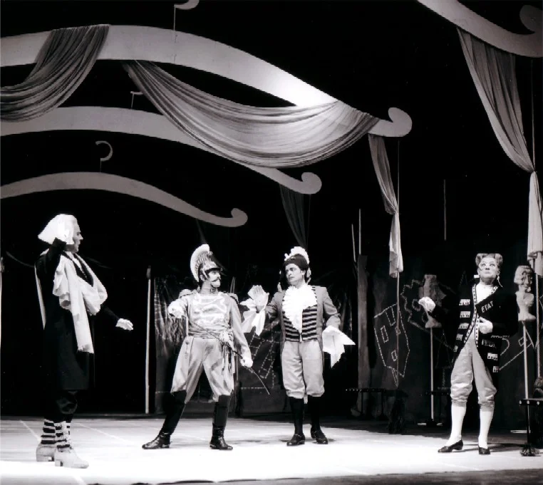 Photo en noir et blanc prise lors de la création de l'opéra de chambre de Bertold Hummel « Les habits neufs de l'empereur », op. 10 (19 juin 1957, Fribourg) : quatre interprètes en costumes de cour discutent sur scène (1re photo)