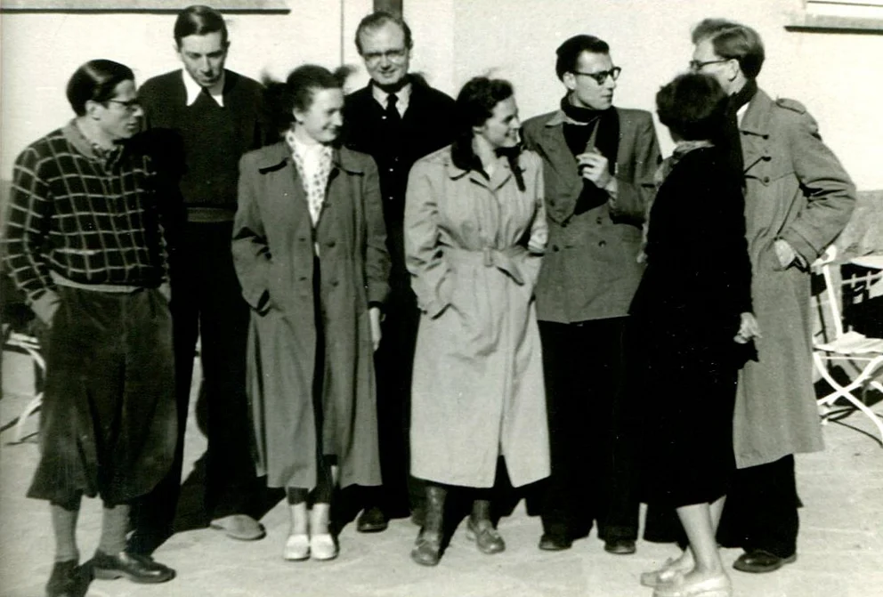 Schwarzweißfoto einer Gruppe von acht Personen in Mänteln, die im Freien beieinanderstehen und sich unterhalten (Uraufführungsinterpreten von Bertold Hummels op. 6)