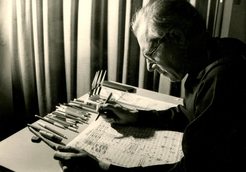 Bertold Hummel composing at a desk covered with sheets of music and pens, 1973.