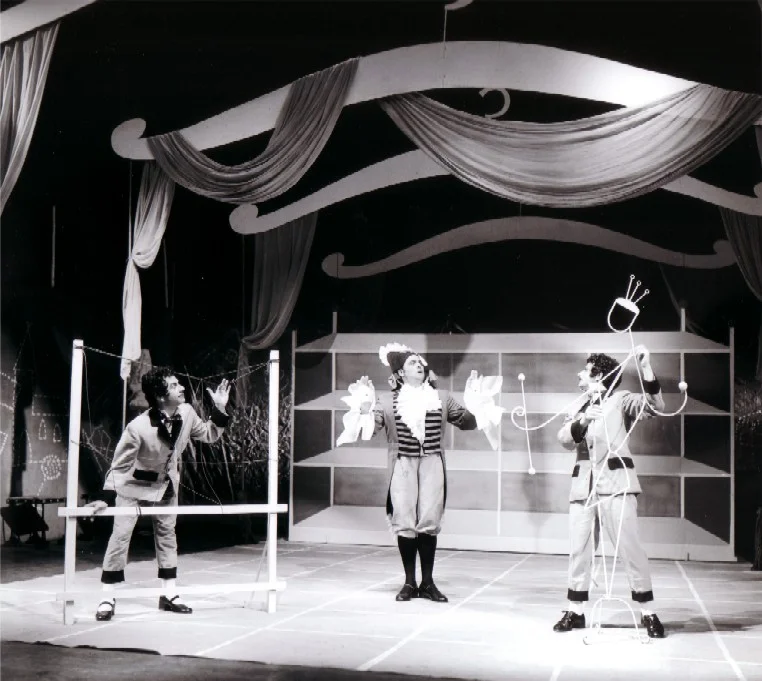 Photo en noir et blanc prise lors de la création de l’opéra de chambre de Bertold Hummel « Les habits neufs de l’empereur », op. 10 (19 juin 1957, Fribourg) : les escrocs déguisés en tisserands examinent, les mains levées, le tissu prétendument invisible ; à gauche, un courtisan devant un châssis tendu de fils (2e image)