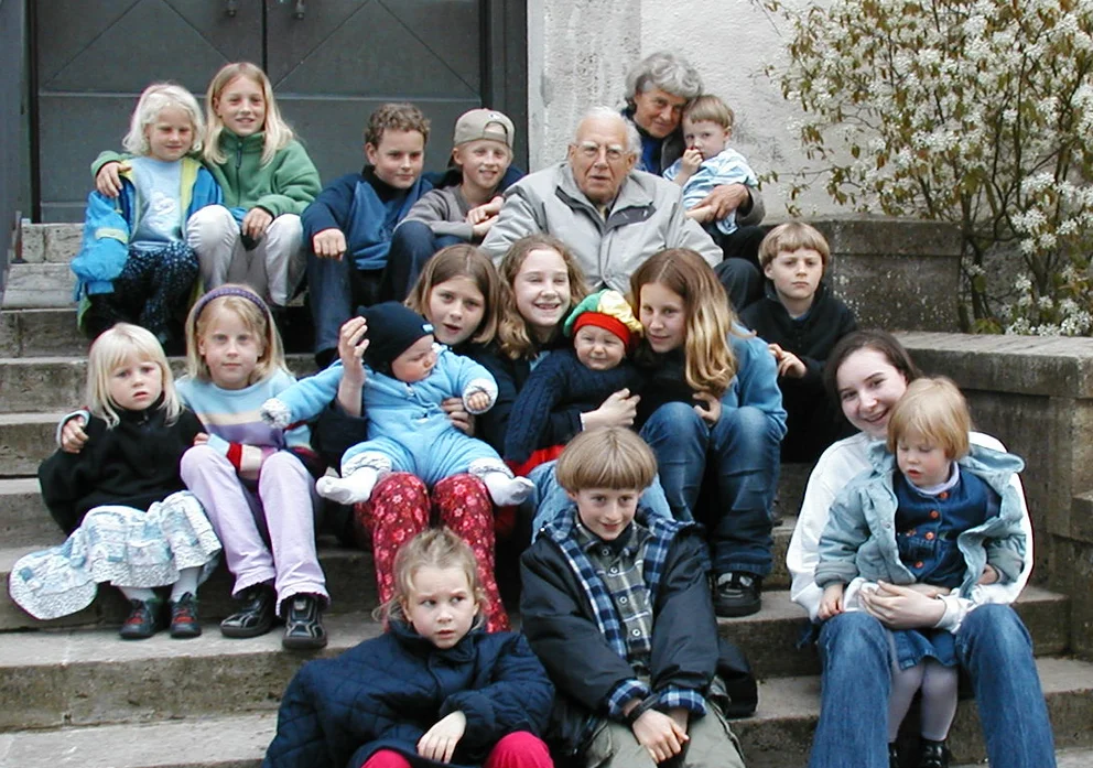 Farbfoto: der Komponist Bertold Hummel und seine Frau Inken sitzen inmitten einer großen Gruppe von Enkelkindern auf einer Freitreppe vor einem Portal, April 2002