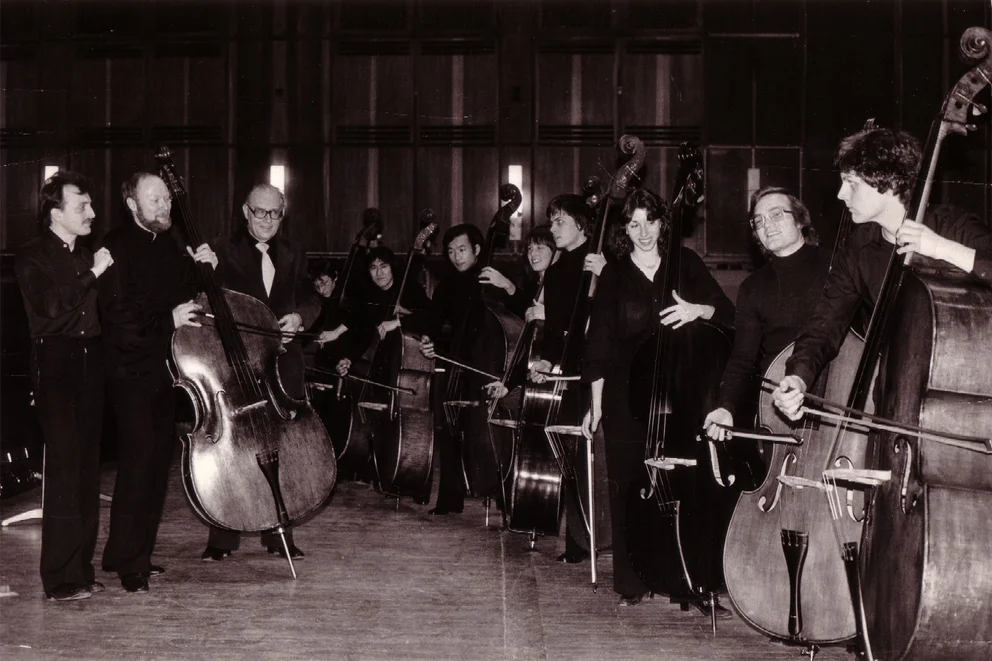 Photo en noir et blanc d'un groupe de contrebassistes avec leurs instruments, rassemblés autour d'un homme d'un certain âge en costume (contexte visuel de la « Sinfonia piccola » op. 66 de Bertold Hummel)