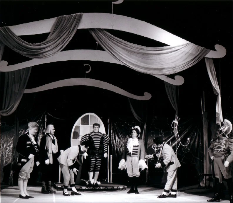 Photo en noir et blanc prise lors de la création de l'opéra de chambre de Bertold Hummel « Les habits neufs de l'empereur », op. 10 (19 juin 1957, Fribourg) : l’Empereur sur un petit podium, entouré de courtisans qui s’inclinent et s’affairent autour de son nouveau vêtement (4e image)